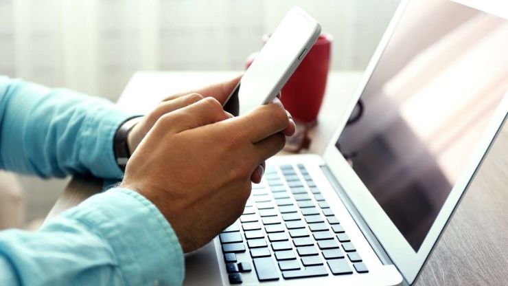 Vīrietis, sēžot pie datora, telefonā meklē kontaktinformāciju savam refinansēšanas kreditoram