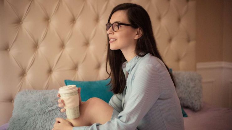 Sieviete aizdomājusies sēž uz dīvāna ar rīta kafijas krūzi un atkārto pozitīvas naudas afirmācijas