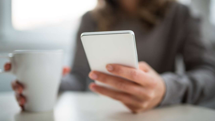 Женщина сидит за столом и читает о разновидностях кредитов на смартфоне, держа в руке чашку с напитком