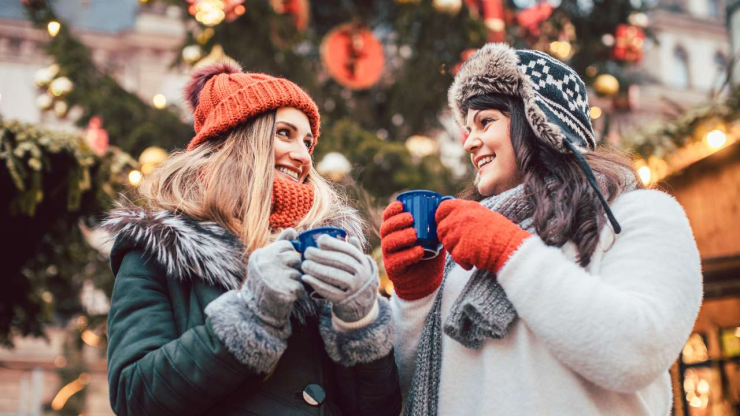 Две подружки у елки на улице пьют горячий напиток и радостно улыбаются – Рождество в самом разгаре