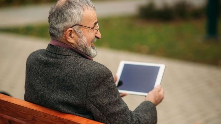 Мужчина сидит в парке на скамейке и ищет в планшете доступный кредит пенсионерам в Латвии