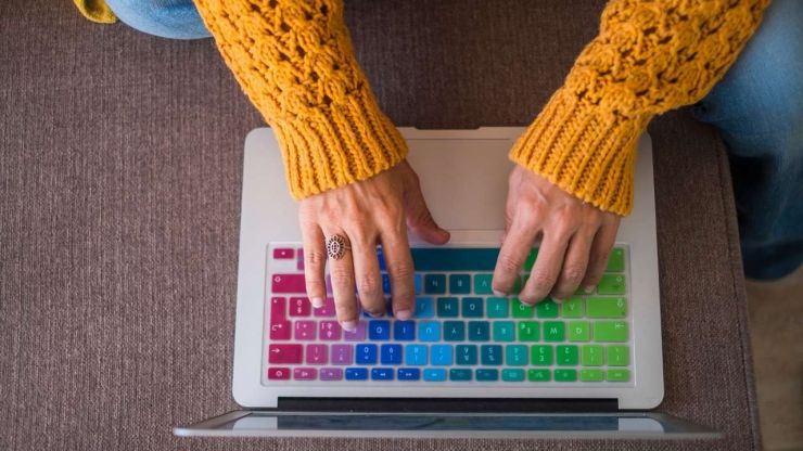 Sieviete dzeltenajā džemperī meklē uz sava datora ar krāsaino klaviatūru, kur var iegūt peļņu internetā