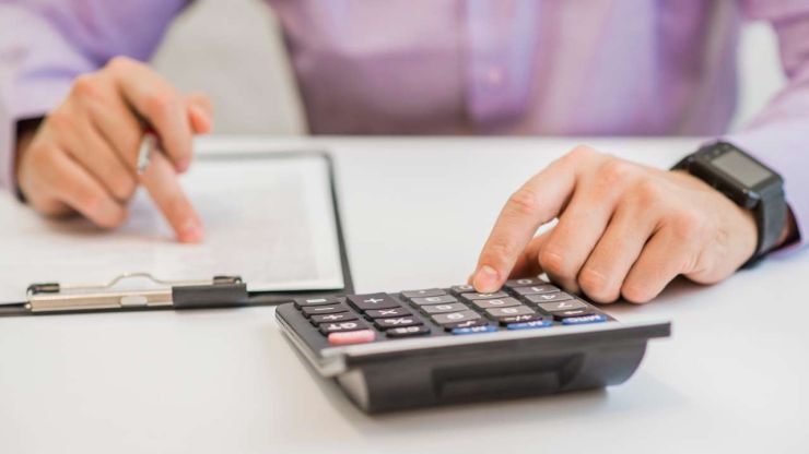 Vīrietis sēž pie galda, lasa kredītlīgumu un aprēķina uz kalkulatora kopējos procentus par aizdevuma lietošanu
