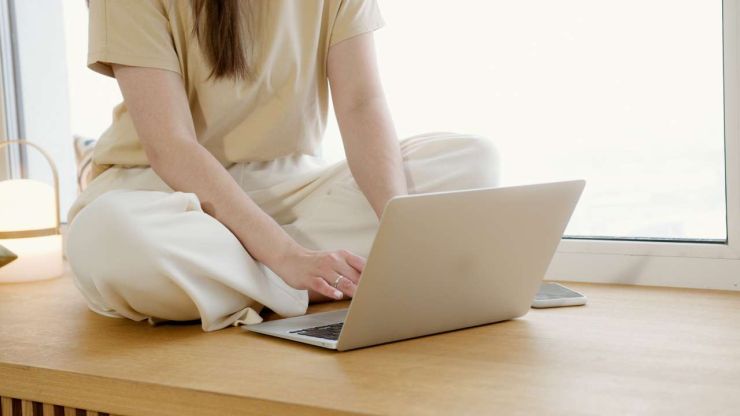 Sieviete sēž uz galda pie loga un portatīvajā datorā meklē, kur paņemt kredītu ar zemu procentu likmi