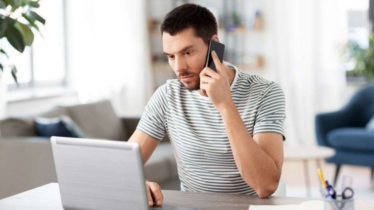 Vīrietis skatās datorā un tur pie auss mobilo telefonu, jo vēlas parunāt ar kreditoru par savu aizdevumu pret ķīlu