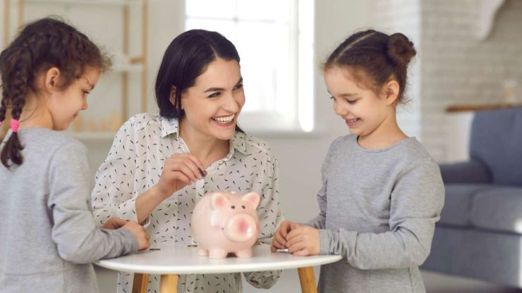 Uz galda stāv krājkasīte, kurā māte ar divām meitām liek monētas, jo apgūst finanšu pratību