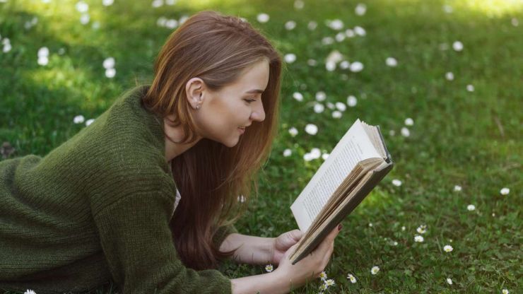 Девушка в парке лежит на траве и читает одну из полезных книг о финансах