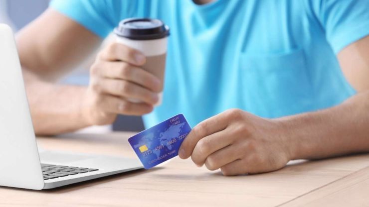 Vīrietis sēž pie datora ar kafijas krūzi un kredītkarti, kurā ir ieskaitīts kredīts uz gadu
