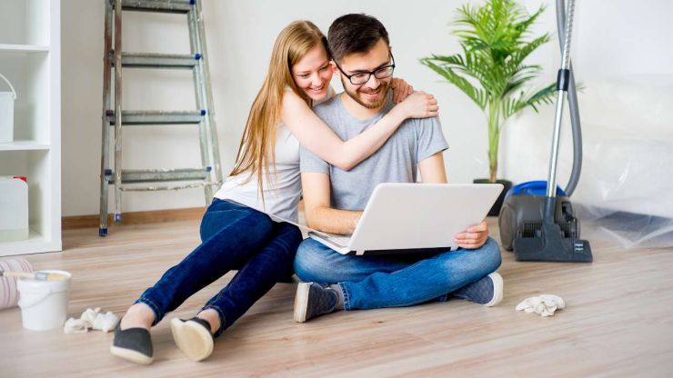 Молодая пара сидит на полу и в ноутбуке смотрят расходы на ремонт квартиры, а на фоне стоит стремянка и пылесос