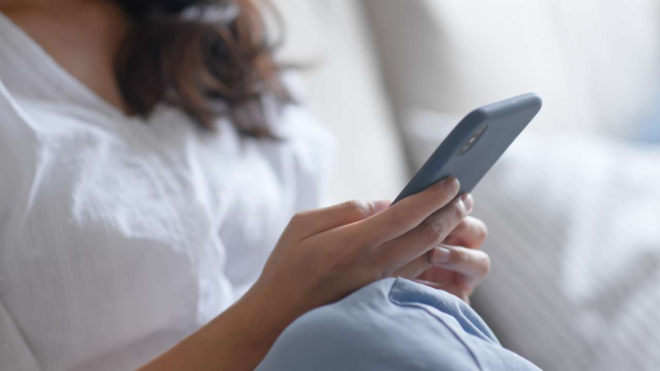Женщина в белой рубашке, сидя на диване, читает в мобильном телефоне основные требования для оформления кредита