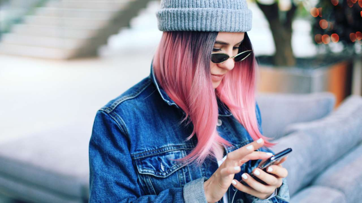 Sieviete ar rozā matiem un džinsa jaku lieto viedtālruni, jo meklē informāciju, kā izmantot mobilo banku droši un efektīvi