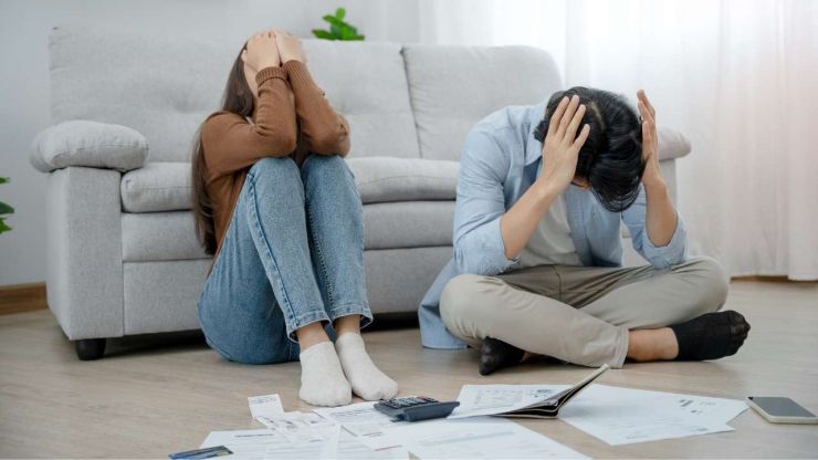 Vīrietis ar sievieti sēž uz grīdas pie dīvāna ar izliktiem parādu rēķiniem, jo viņiem ir atkarība no ātrajiem kredītiem