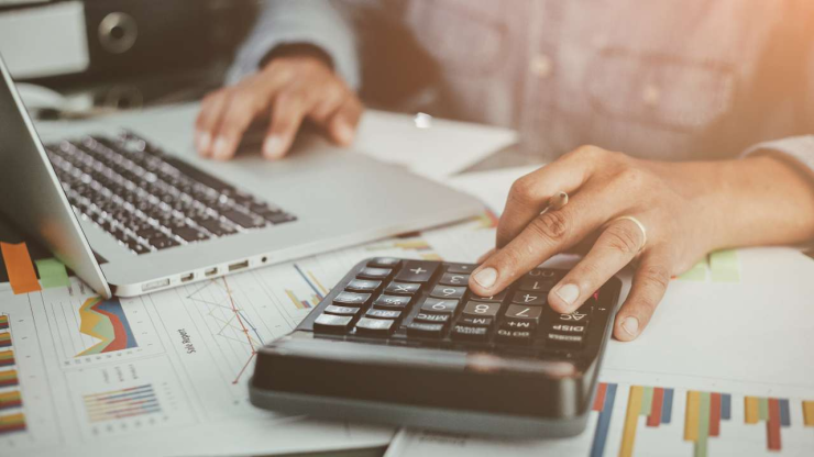 Cilvēks sēž pie galda ar datoru, dokumentiem un uz kalkulatora rēķina nepieciešamo finansējumu savam biznesam