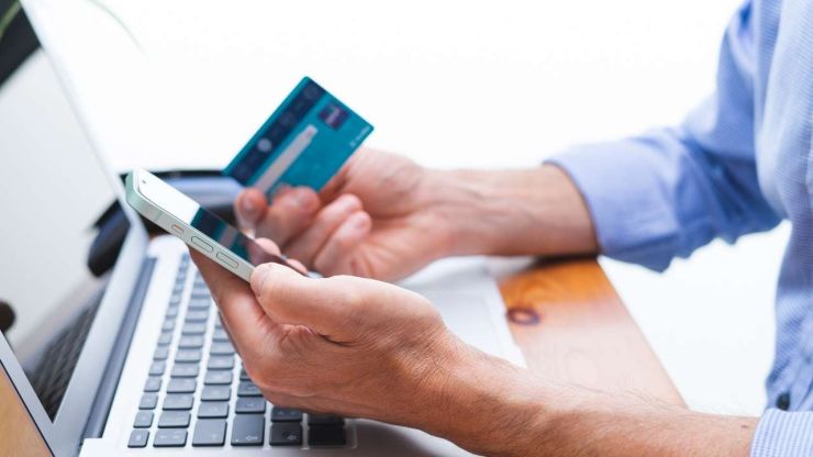 Vīrietis, sežot pie datora un turot kredītkarti, meklē telefonā interneta kredītu