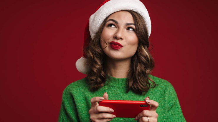 Sieviete rūķa cepurē un zaļajā džemperī viltīgi smaida, jo izvēlējās dāvanām svētku kredītu, kuru tagad noformē telefonā