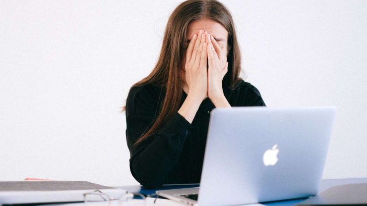 Sieviete, aizsegusi seju ar plaukstām, sēž pie atvērta datora un domā, ka aizdevumi ir negatīvi ietekmējuši viņas dzīvi
