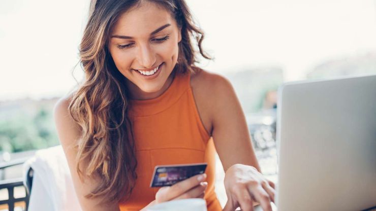 Девушка сидит за ноутбуком и улыбается, потому что делает покупки при помощи кредитной карты