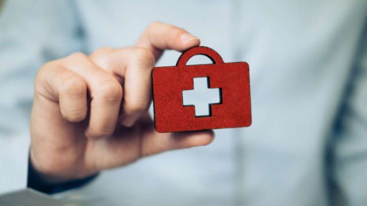 Мужская рука врача держит красную фигурку медицинского чемодана – страховка помогает экономить на медицинских расходах