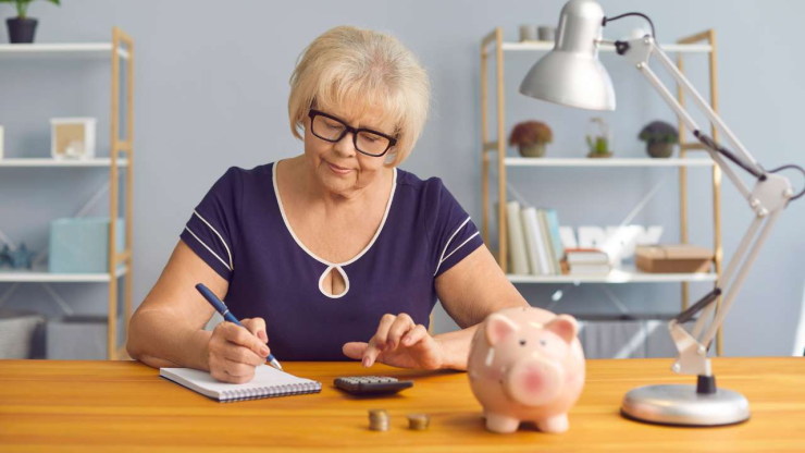 Пожилая женщина сидит за письменным столом и рассчитывает размер 1 пенсионного уровня после индексации