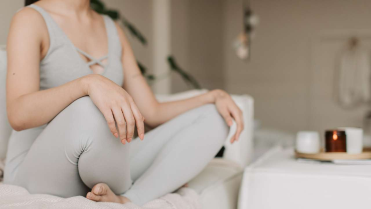 Sieviete mājīgā atmosfērā sēž uz dīvāna ar saliktām kājām, jo meditācija veicina mentālo veselību