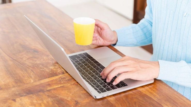 Persona zilā džemperī ar dzelteno kafijas krūzi rokā meklē datorā, kur ir pieejami  kredīti bez darba vietas