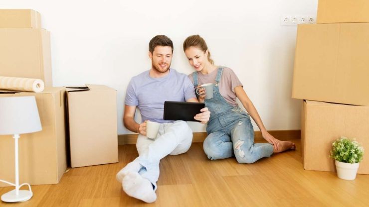 Молодая пара сидит на полу среди коробок в своем новом доме, который приобрела благодаря ипотечному кредиту, и рассматривает в планшете проект