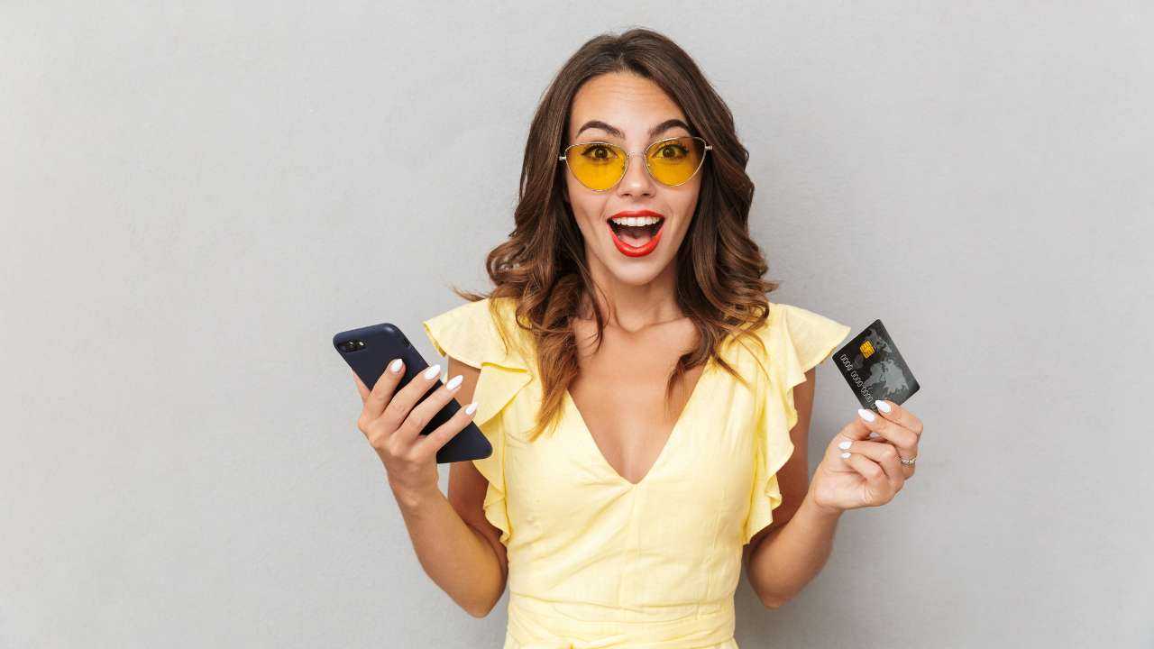 Девушка в одной руке держит телефон, в другой – банковскую карточку, и радуется, потому что получила одобрение на СМС кредит