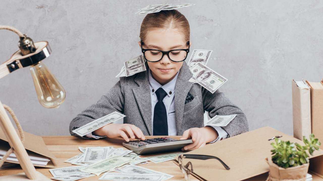 Маленькая «бизнес-леди» считает на калькуляторе свои карманные деньги, которые лежат вокруг нее на столе, ее плечах и голове
