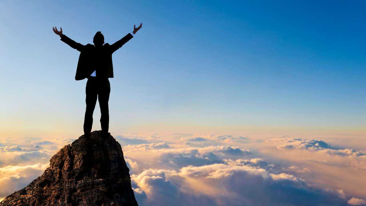 Финансово успешный мужчина в костюме стоит на вершине горы, потому что достигает своих целей