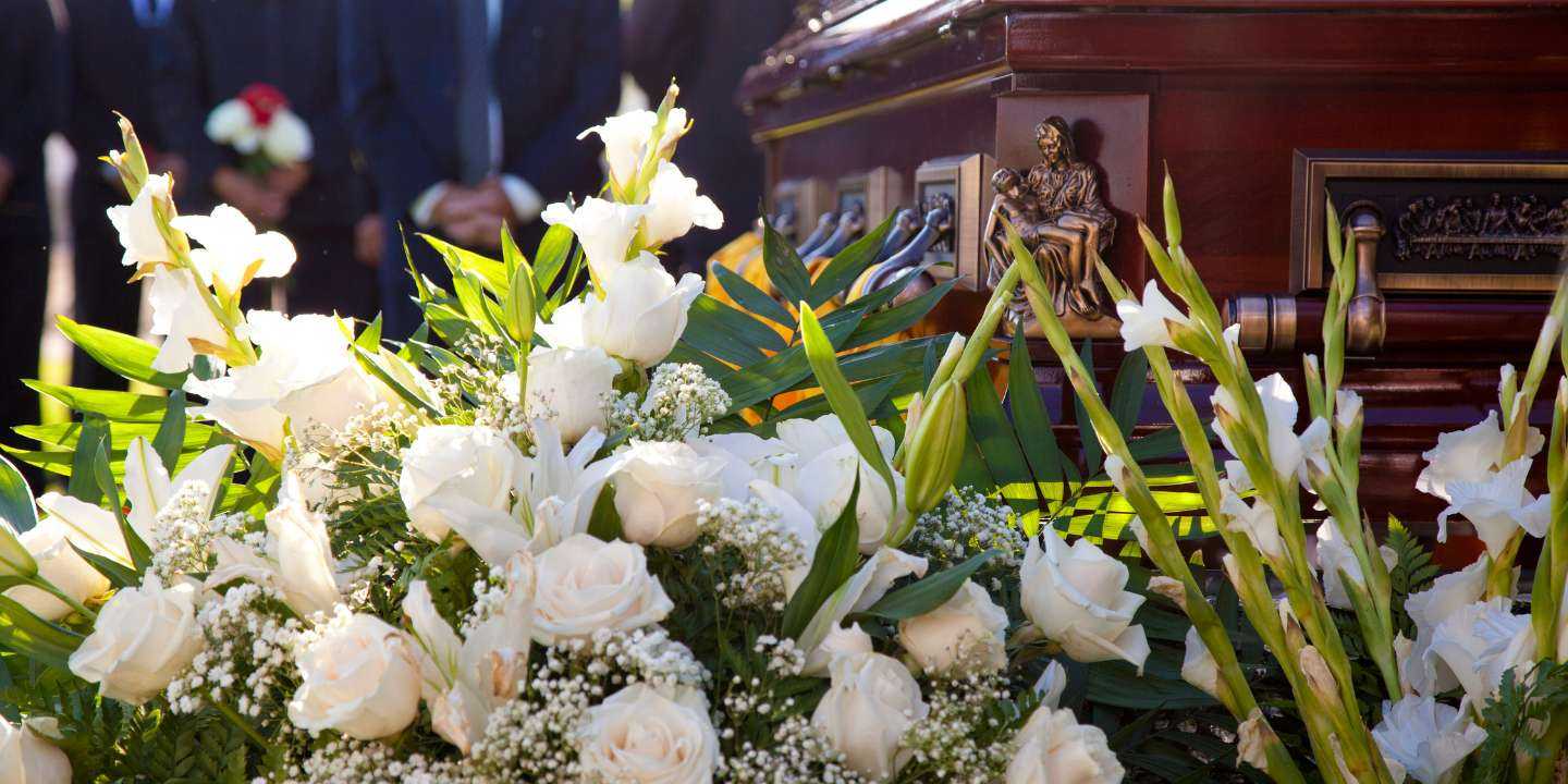 Priekšplānā balto ziedu pušķis un tumšā koka zārks bēru ceremonijā, ko norīkoja cilvēks, kas saņēma apbedīšanas pabalstus