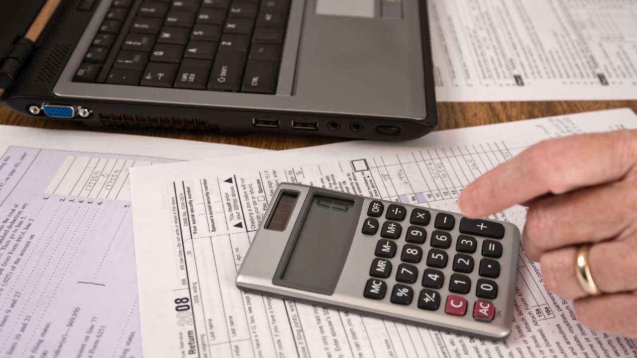 Предприниматель сидит за столом и на калькуляторе рассчитывает налог на прибыль в Латвии, который ему необходимо заплатить