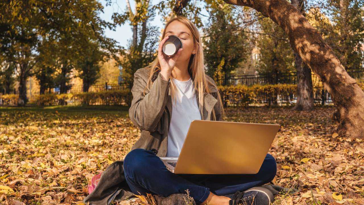 Sievietē sēž parkā uz zāles un rudens lapām, dzer kafiju un datorā izstrādā jaunu partnermārketinga stratēģiju