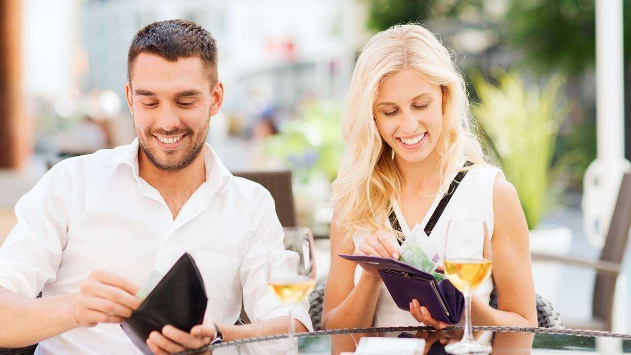 Vīrietis un sieviete, sēžot pie galdiņa kafejnīcā, smaida, vērot vaļā maku ar darba samaksu par papildu veikto darbu