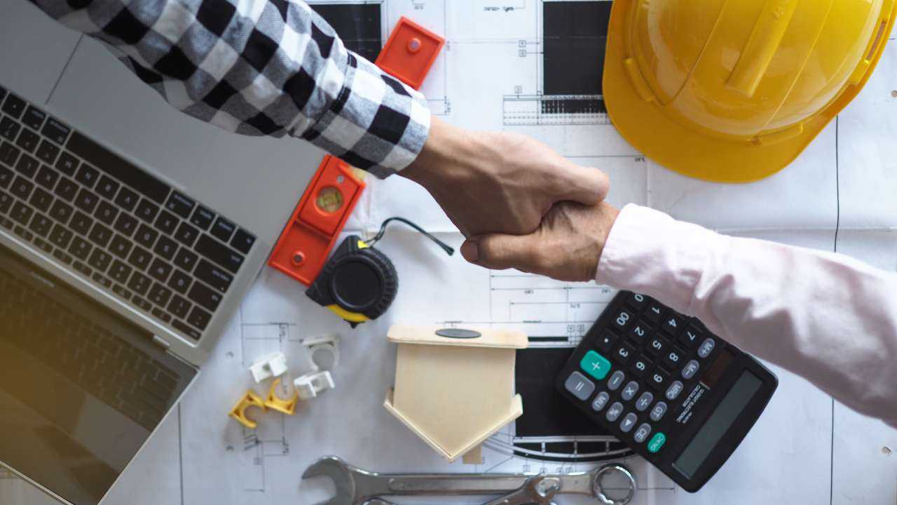 Uz galda izvietoti dators, celtniecības instrumenti, un vīrieši spiež rokas, jo ir vienojušies par mājokļa uzlabošanas projektu