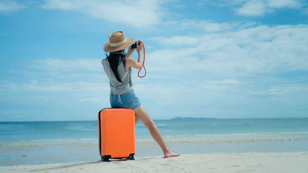 Девушка в соломенной шляпке действует по плану своего путешествия, поэтому сидит на оранжевом чемодане на берегу моря и фотографирует