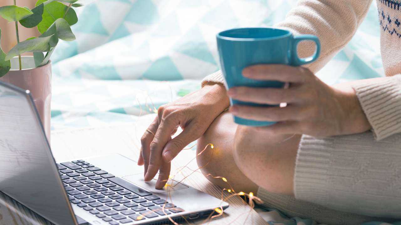 Девушка сидит на кровати с кружкой чая и в ноутбуке сравнивает финансовые компании, чтобы выбрать кредит