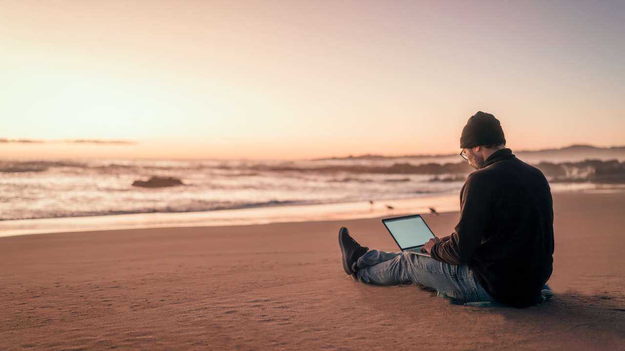 Мужчина сидит на песке на берегу моря и через ноутбук зарабатывает при помощи партнерского маркетинга