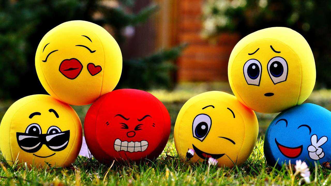 На траве шесть разноцветных мягких мячиков с выражениями разных эмоций, которые могут влиять на финансовые решения