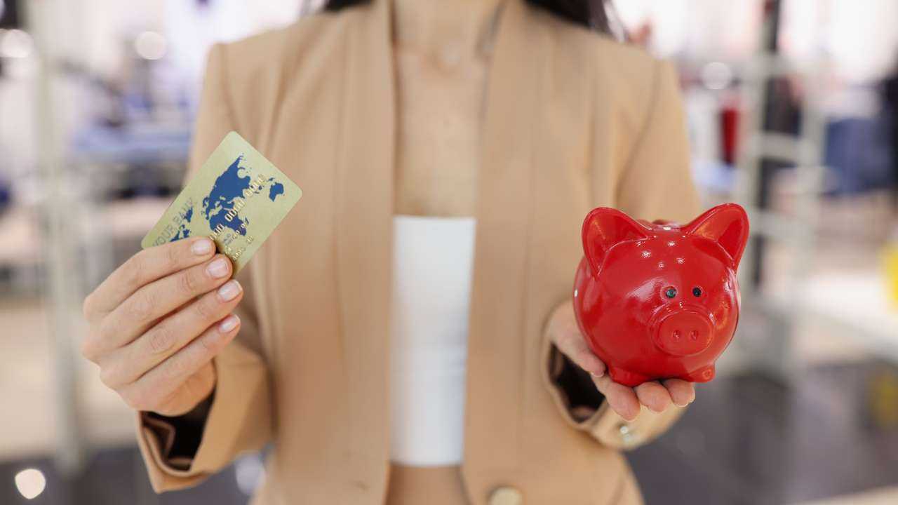 Девушка стоит и держит в одной руке расчетную карту, а в другой красную свинку-копилку, потому что решает, какой способ накопления денег использовать