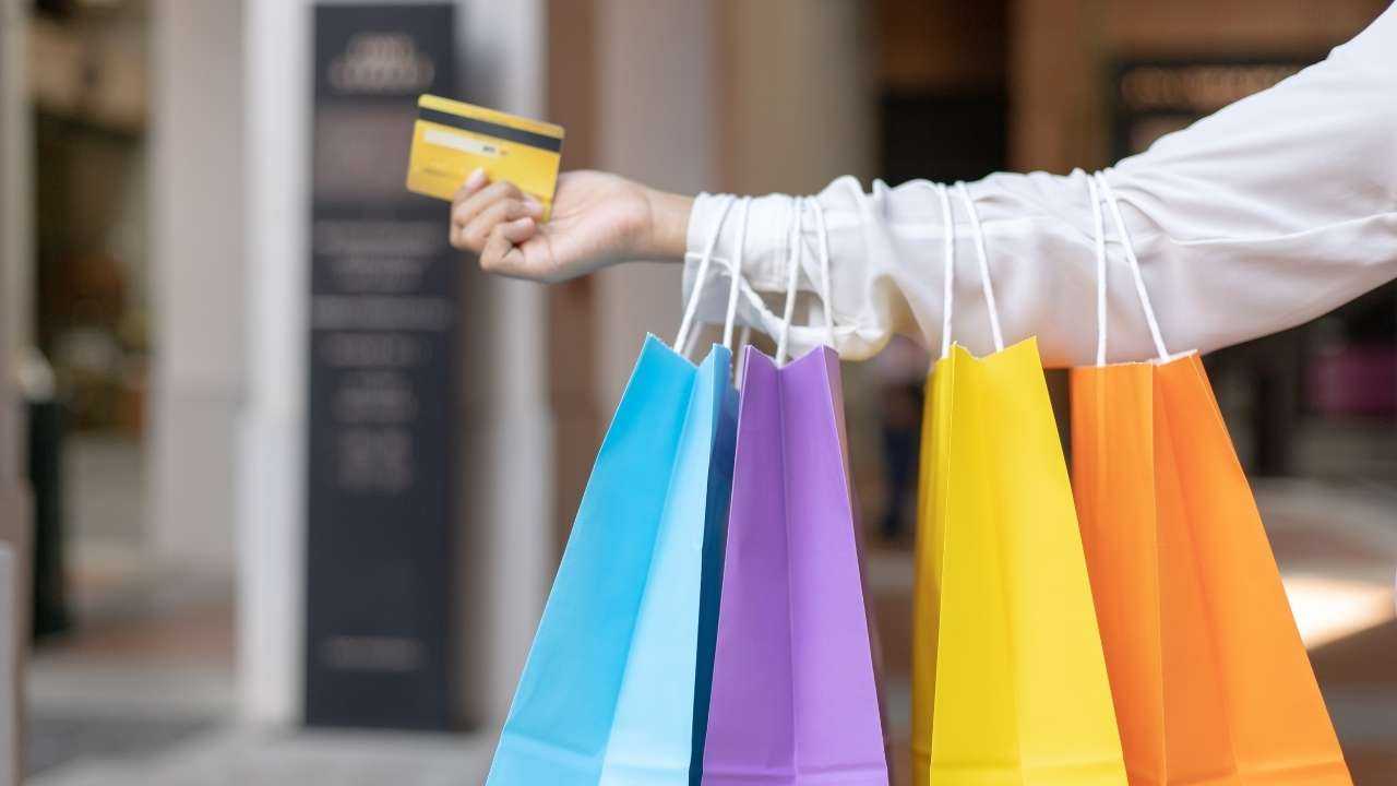 Persona baltā kreklā tur rokā kredītkarti ar kredītlīniju un četrus krāsainus papīra iepirkumu maisiņus