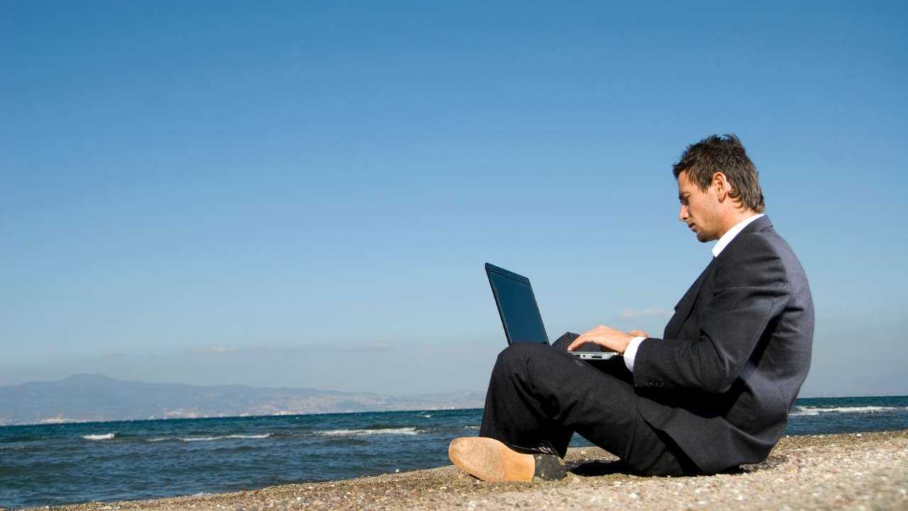 Мужчина с ноутбуком сидит на берегу моря и обдумывает возможность оформить кредит для бизнеса