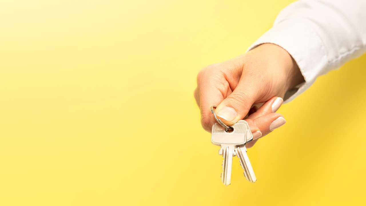 Dzeltens fons un sieviete baltā kreklā sniedz pircējam mājas atslēgas