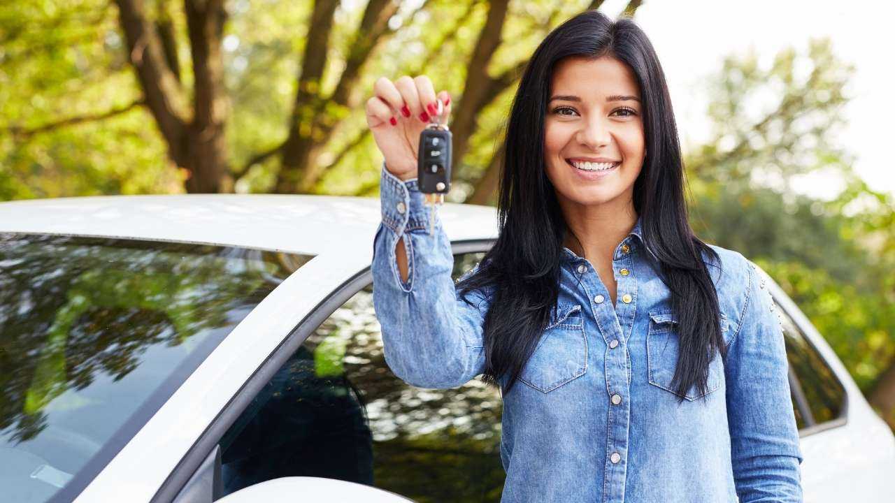 Девушка стоит на фоне машины, которую купила благодаря кредиту на покупку авто, и улыбаясь показывает ключ от новой машины